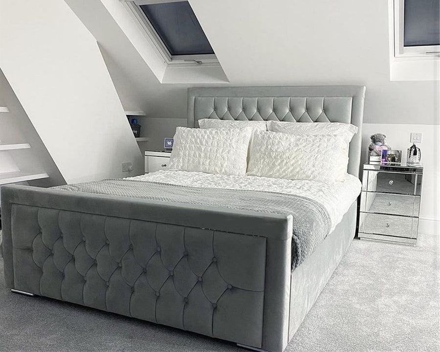 A modern bedroom with a tufted grey upholstered bed frame, white bedding, and a mirrored nightstand under a slanted ceiling with a skylight.